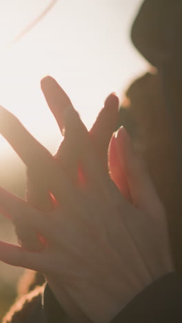 fotografía de cerca de una persona frotando las manos cerca de su pecho, perdida en sus pensamientos. la cara no es visible, pero la persona lleva un abrigo marrón y una sudadera con capucha. luz del sol en el fondo