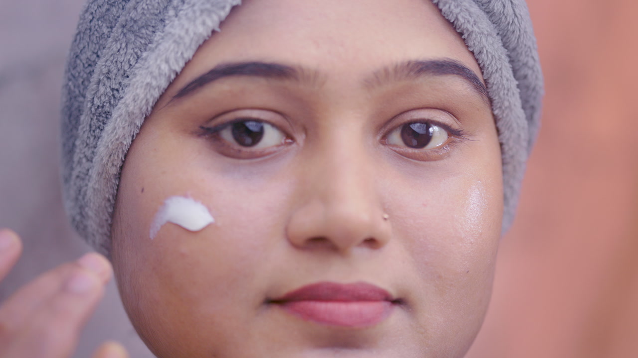 Close up shot of Indian woman gently massaging moisturizing cream on her cheeks in modern bathroom.