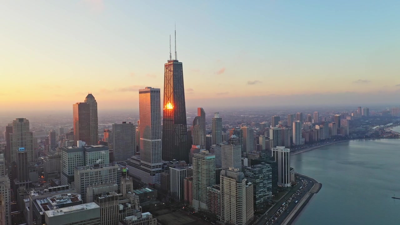 Filmagem aérea fluida movendo-se para a direita sobre Chicago ao pôr do sol, exibindo a 875 North Michigan Avenue e o horizonte circundante da orla do lago brilhando com a luz quente do entardecer ao lado do Lago Michigan.