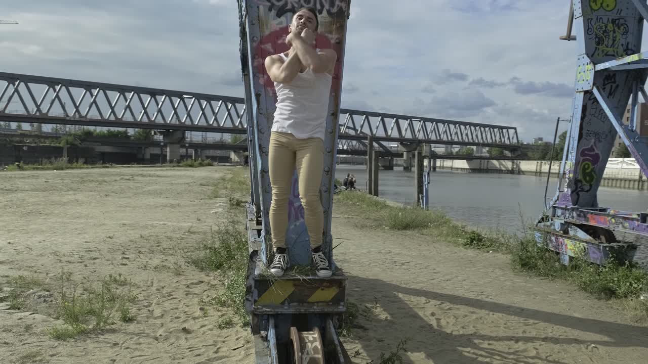 Pan right view of modern hipster male in casual wear standing on metal platform of abandoned crane with graffiti and performing dynamic dance movements with arms