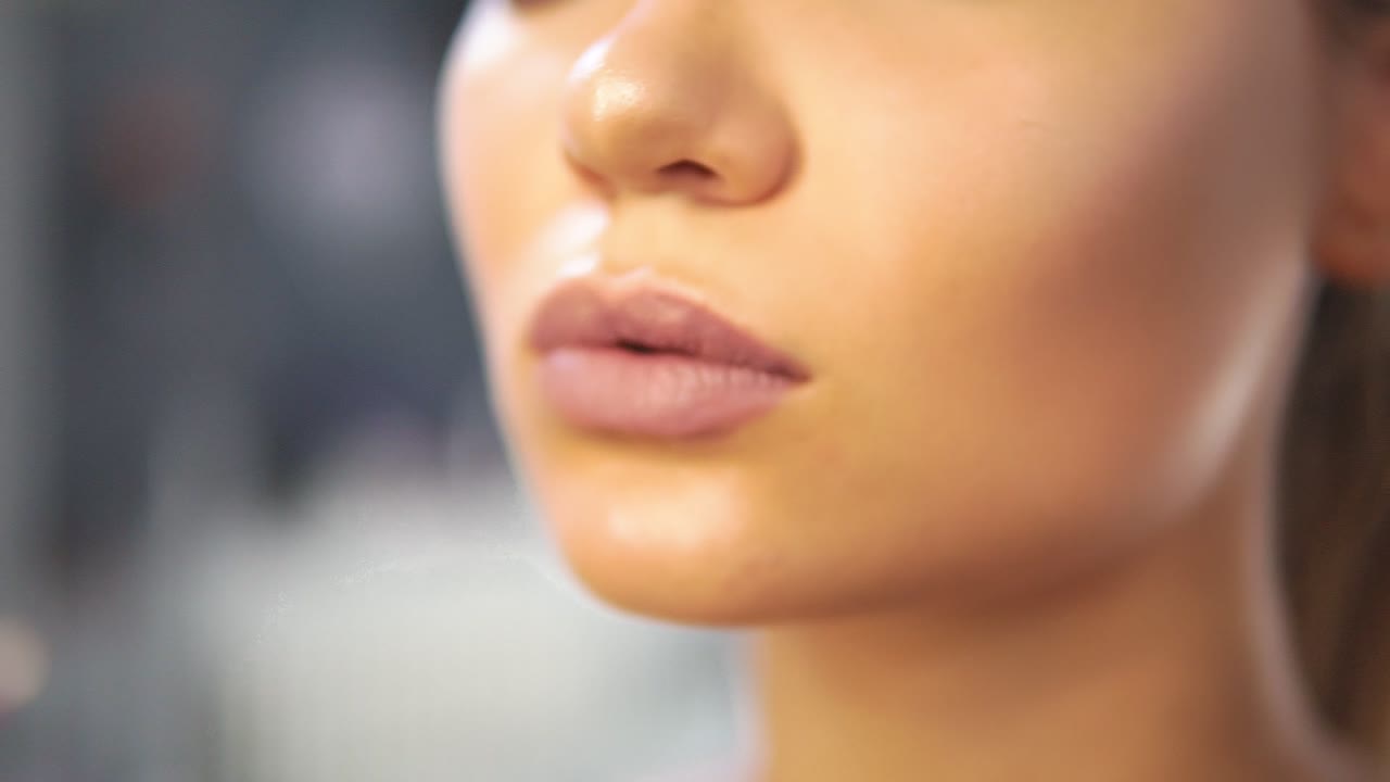 Close Up view of a brush with golden sparkle gloss applying professional makeup on the model's lips. Lip gloss on the lips