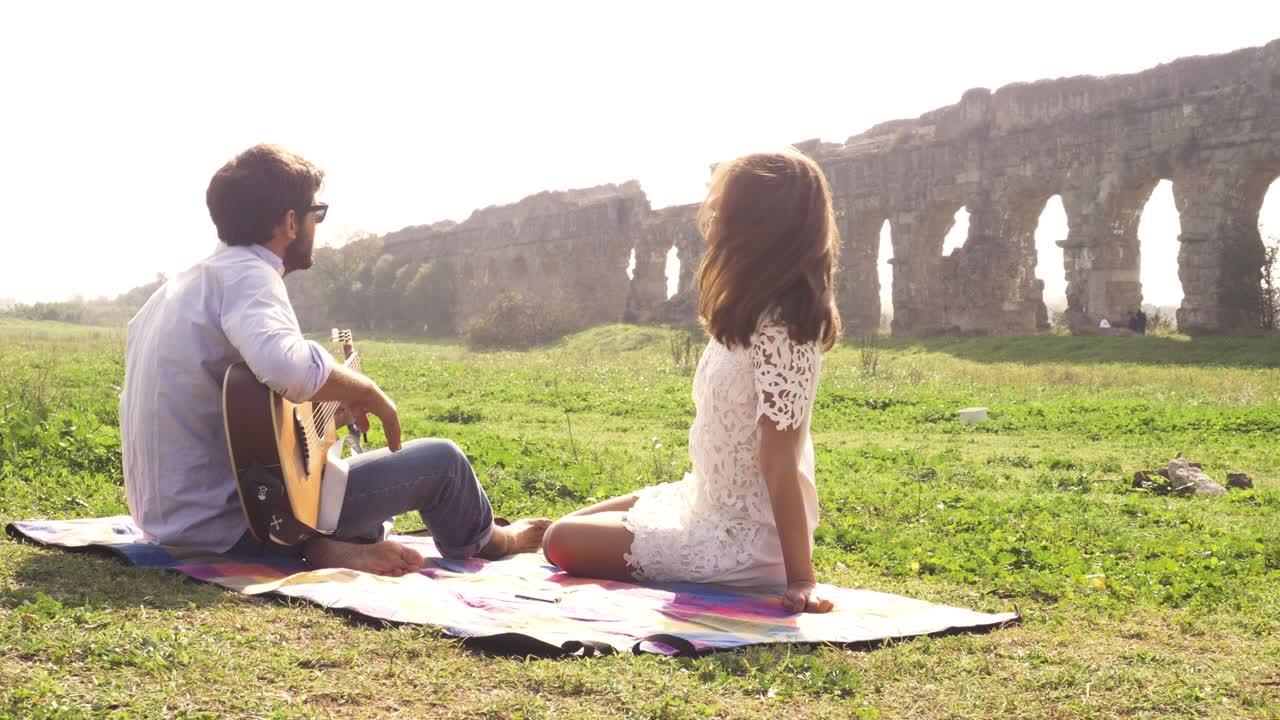 행복한 젊은 사랑스러운 부부는 잔디에 담요 위에 누워 앉아 로마의 고대 아쿠에듀크 앞에 있습니다. 로마의 파르코 데글리 아쿠에두티 공원의 폐허. 로맨틱한 웃음. 기타를 연주하고 노래합니다. 아름다운 소녀. 모자를 던집니다.