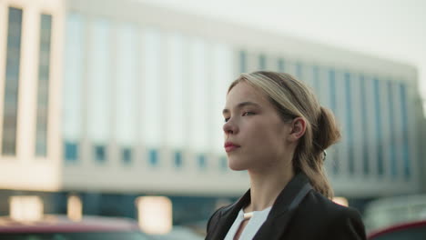 Close up of professional lady in black suit smiling slightly with confident expression and blurred background of modern building and cars in urban environment