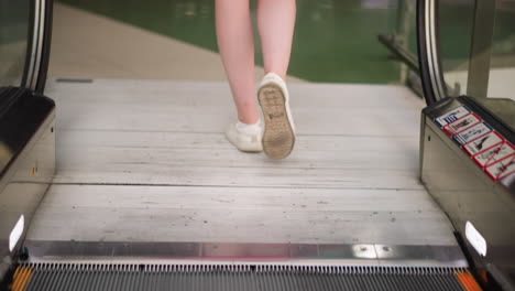 leg view of a white lady wearing white canvas sneakers as she steps out of a moving escalator, her reflection is visible on the glass frame of the escalator, and signs are visible on the escalator