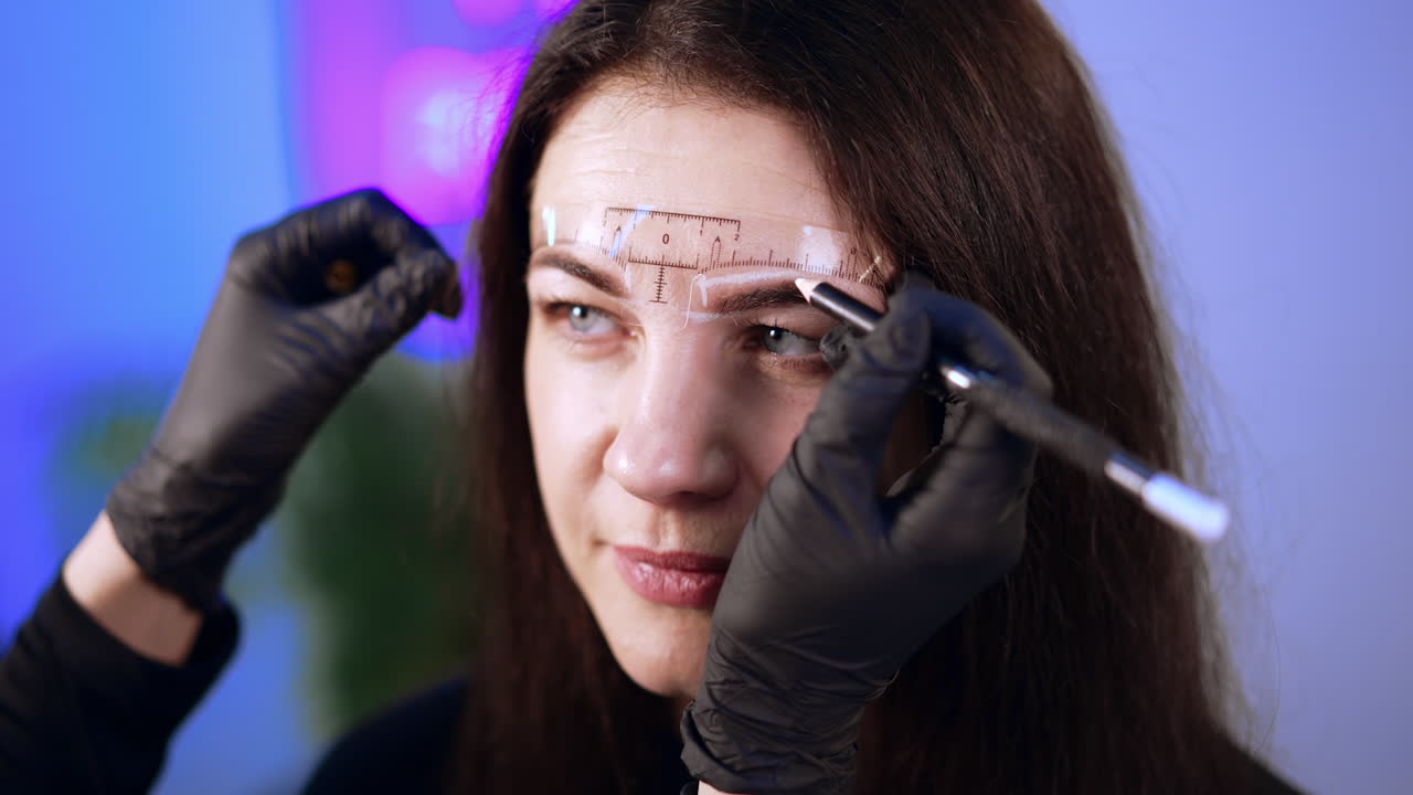 Black gloved hands of beautician uses white pen to mark eyebrow lines for the client. Brunette woman is prepared for eyebrow permanent make up.
