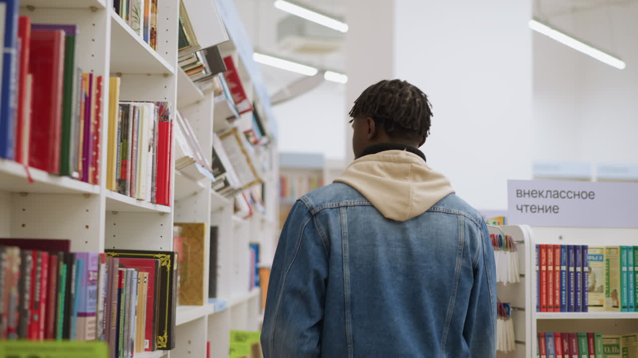 Back view of young person in denim jacket with hoodie, walking down bookstore aisle. Shelves filled with books in various genres, well-organized and brightly lit, with a clear view of colorful book spines