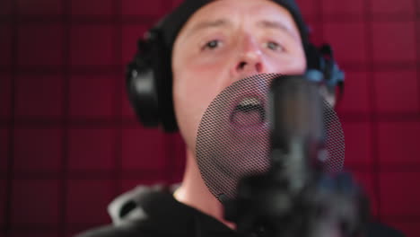 un hombre vestido de negro está usando auriculares y de pie frente a un micrófono en una habitación a prueba de sonido roja. está cantando con seriedad, haciendo un gesto de la mano mientras se centra en su actuación