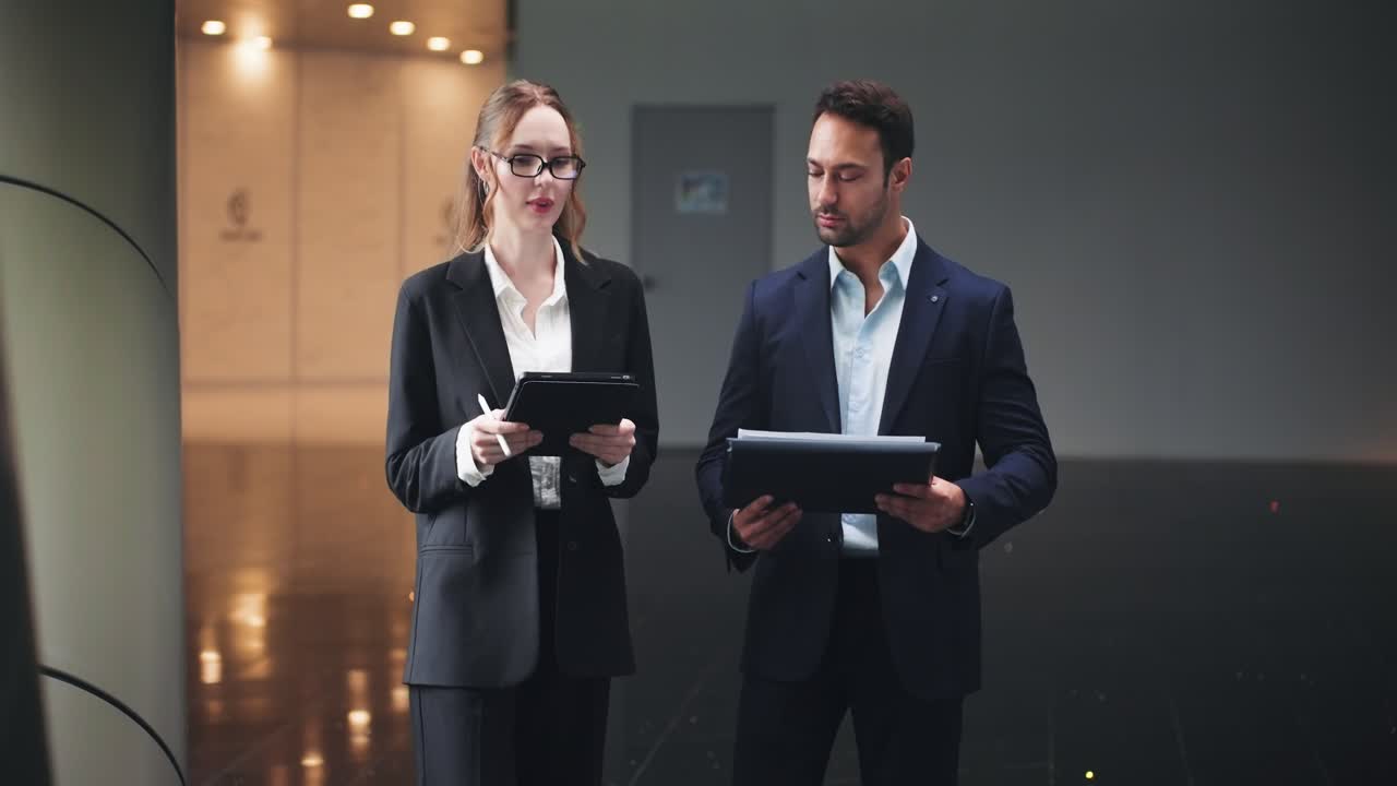 Uomo d'affari europeo che ascolta una donna d'affari con un tablet mentre discutono di un report camminando nel corridoio dell'ufficio. - campo lungo