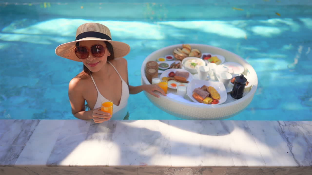 A lovely Asian lady is ready to drink orange juice and eat exotic breakfast from a floating tray in the hotel resort swimming pool. View from the top angle. Slow-motion