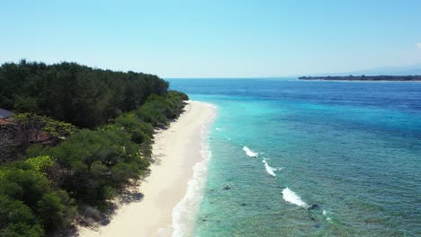 Quiet-white-beach-of-tropical-island-with-lush-vegetation-at-sunrise,-washed-by-white-waves-of-blue-azure-sea-on-a-bright-blue-sky-in-Bali