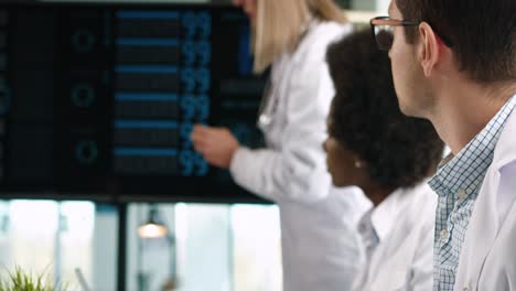 Close-up-view-of-Caucasian-male-doctor-typing-on-tablet-and-looking-at-digital-screen-presentation-sitting-at-meeting-with-multiethnic-doctors-group