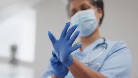 Senior-african-american-female-doctor-wearing-face-mask-putting-protective-gloves-on