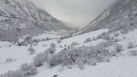 Toma-De-Drones-De-Un-Valle-Lleno-De-Nieve-En-Los-Pirineos-Montañosos-De-España-Con-Una-Pequeña-Casa-Rústica-Al-Final-En-Un-Día-Nevado.