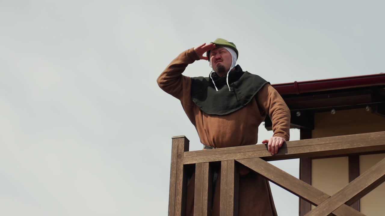 Ein reifer bärtiger Mann mit Metallhelm und gepolstertem braunem Mantel steht auf einer erhöhten hölzernen Aussichtsplattform, hält das Geländer fest, schirmt die Augen ab und scannt den fernen Horizont mit wachsamer Konzentration.
