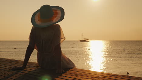 Una-Mujer-Joven-Se-Encuentra-Con-El-Amanecer-En-El-Muelle,-Se-Sienta-Y-Mira-El-Sol-Y-El-Barco-En-El-Mar-Dre