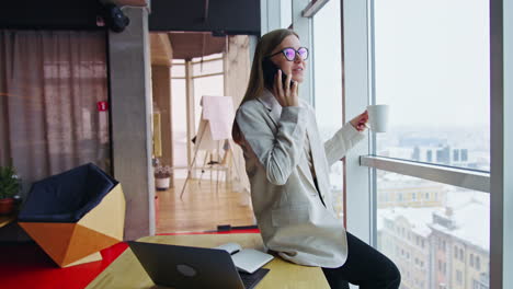 resilient lady sits on the desk, speaks on the phone and holds a cup. lunch break in office with big windows. cityscape at backdrop.