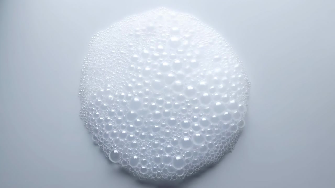 Vista dall'alto centrata di un disco di schiuma bianca densa in evidente cambiamento, con l'area superiore che forma una micro-schiuma fine mentre il bordo inferiore rivela bolle traslucide più grandi su uno sfondo chiaro e pulito