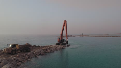 long reach excavator machine receives stones to build a stone pier in the sea, sunset parallax shot