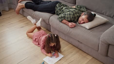 top view of a little brunette girl in a pink dress lies on a wooden floor and draws on a white sheet of paper while her single father a brunette man in a green checkered shirt sleeps on a gray sofa in a modern apartment