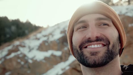 a handsome and cute millennial male bundled in winter gear smiles overlooking an amazing snowy and wintry rocky mountain view