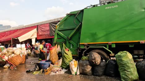 workers gather and load trash onto a green garbage truck.