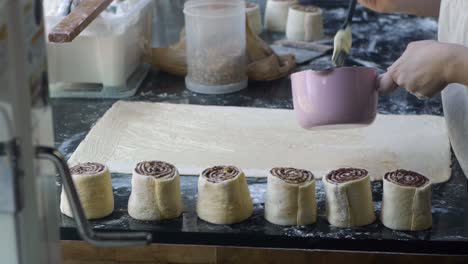 melted butter brushed onto freshly rolled sheet of dough on kitchen table top with cinnamon rolls on display nearby