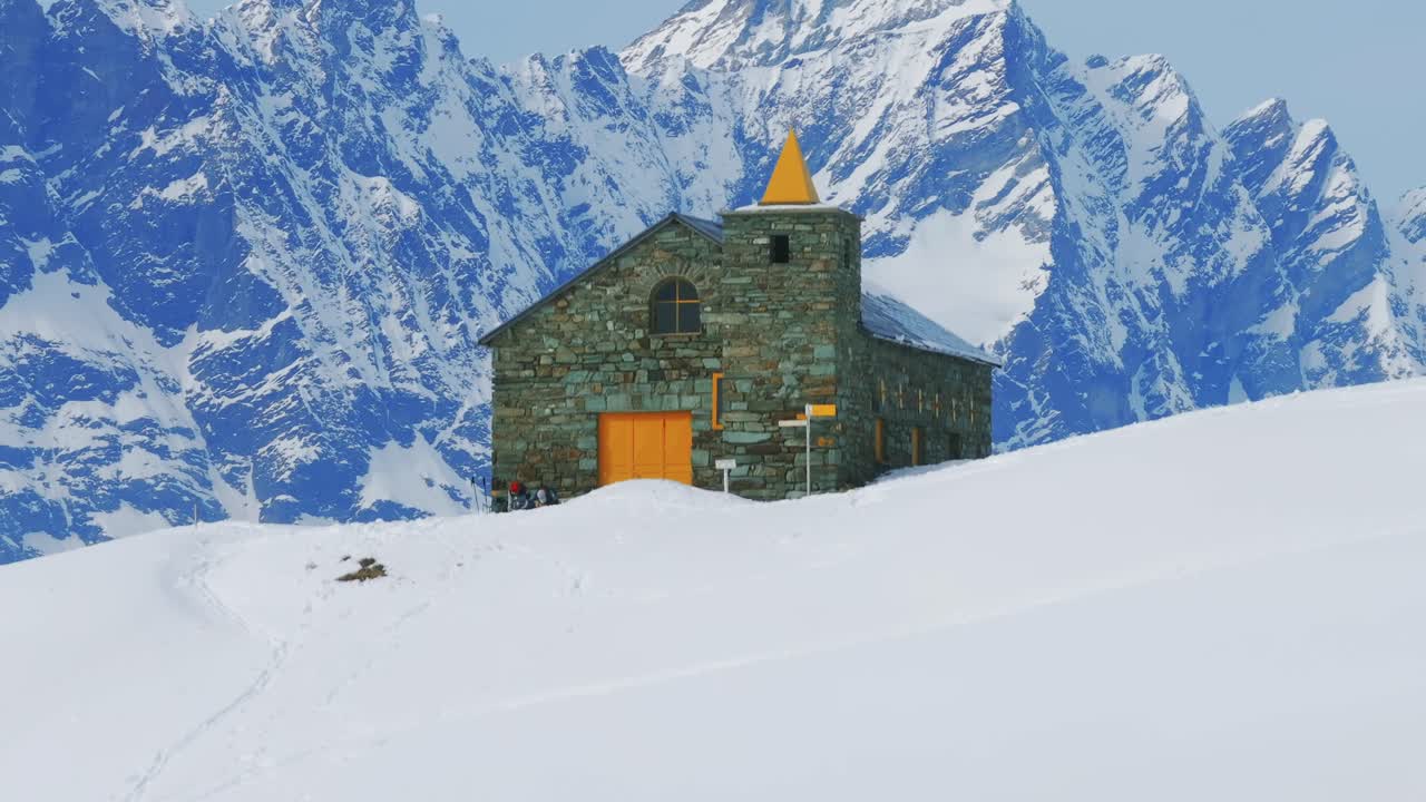 Cappella di San Bernardo chapel with yellow spire, snow-covered mountain backdrop in Cheneil, Aosta Valley, Italy. Aerial drone ascending