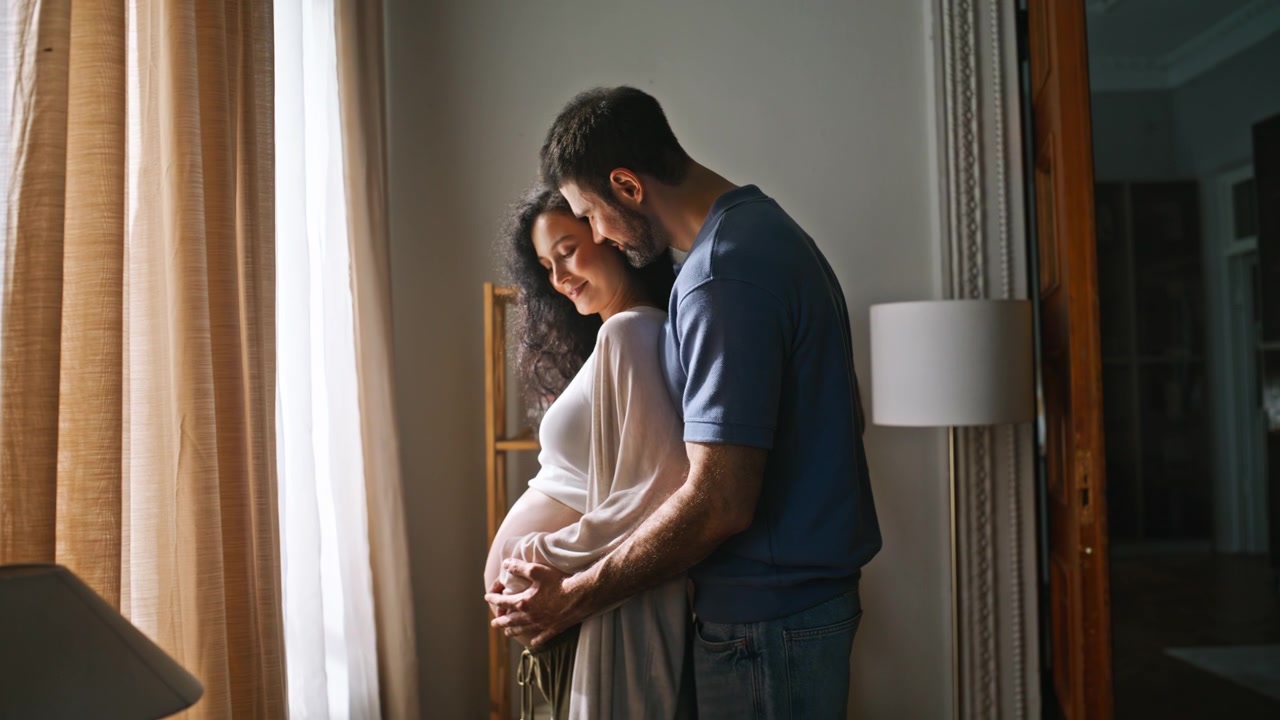 Expecting parents embracing window home. Man holding pregnant belly, maternity.