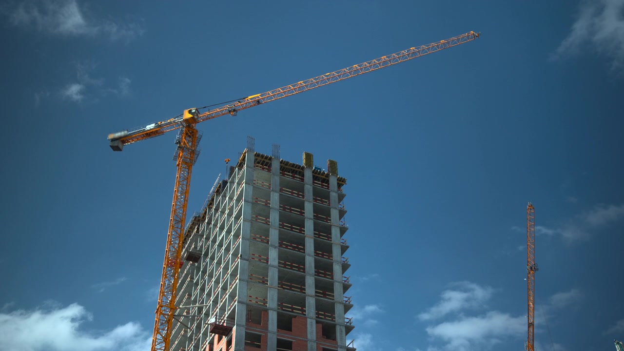 Un alto edificio grattacielo è in costruzione con una grande gru a torre che si estende in alto a sinistra e un'altra gru visibile in lontananza.