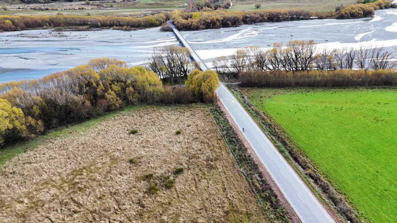 Aerial footage captures a tranquil road flanked by lush fields and a river in Glenorchy, New Zealand