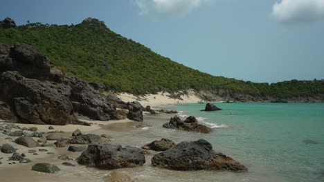 Motion-video-of-waves-hitting-stones-at-shore-of-Colombier-beach-with-green-hills-at-background-in-French-West-Indies,-French-Antilles,-Caribbean-Sea