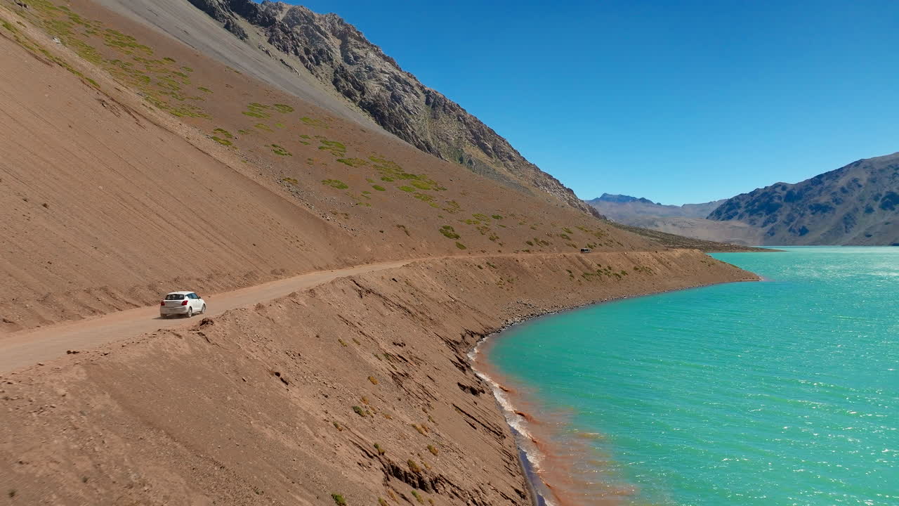 Car driving on dirt road alongside turquoise water of Embalse El Yeso reservoir, rugged Andes mountains under clear blue sky, Chile. Aerial forward