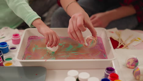 mother and girl roll eggs in tray with paints and oily water