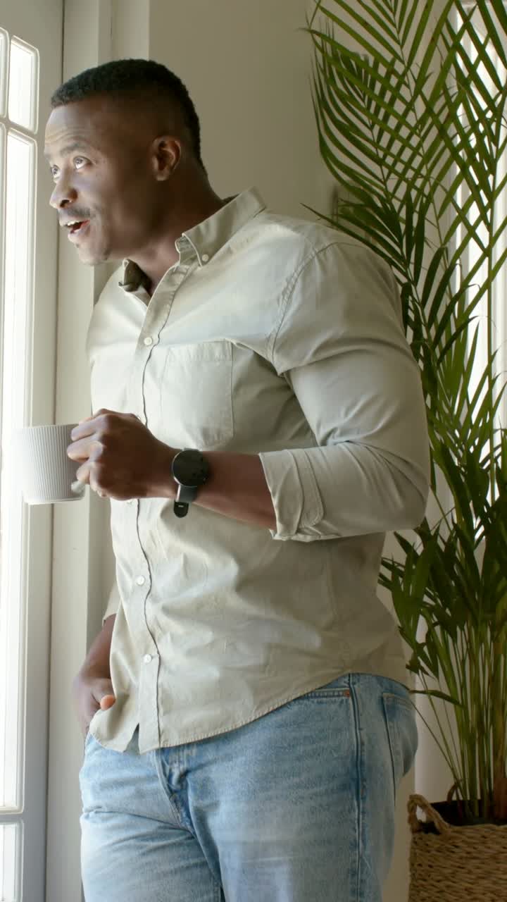 Vertical video: Sipping beige-shirted man lowering white mug, peering at bright window, smiling
