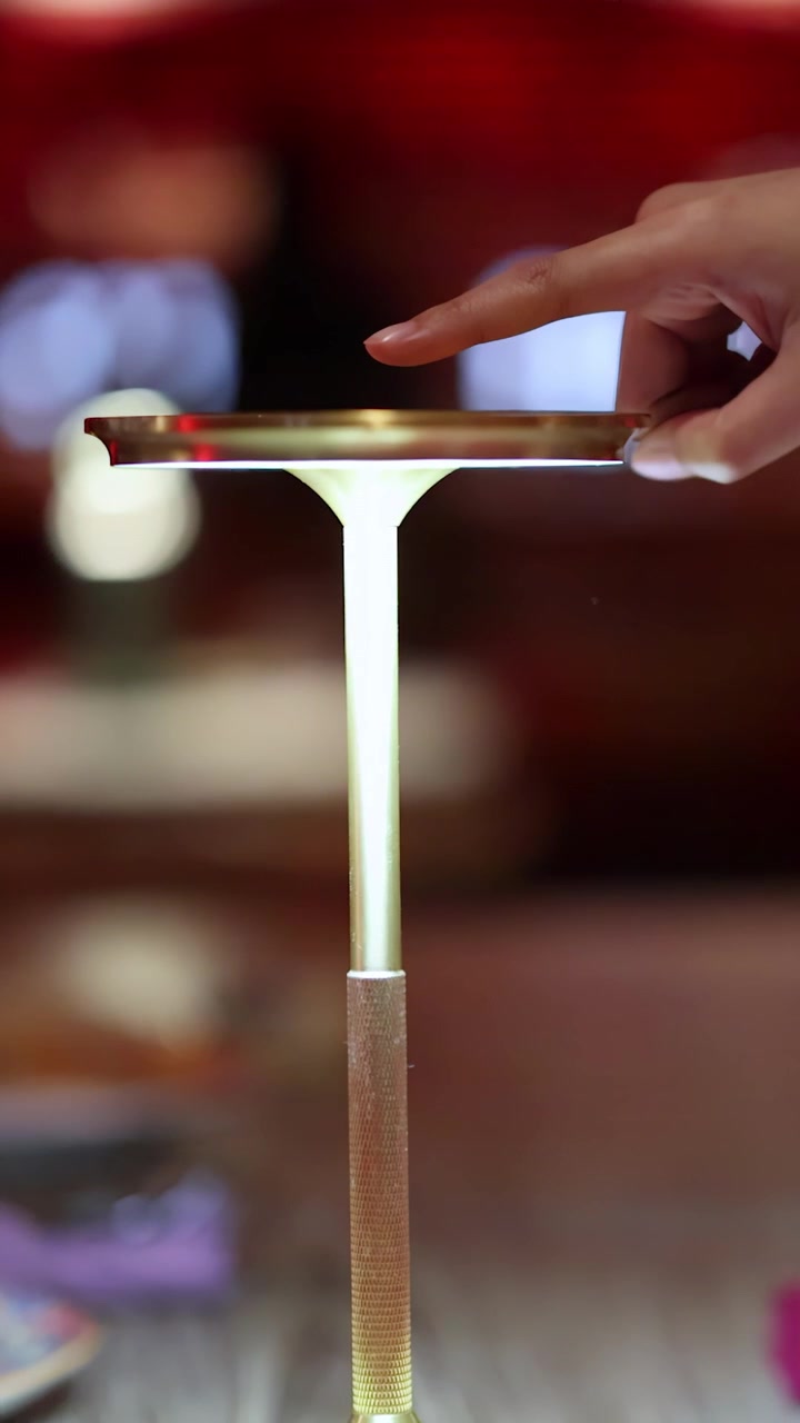Uma mão toca o topo de uma luminária de toque moderna, alternando sua luz em uma mesa de restaurante com iluminação ambiente acolhedora.