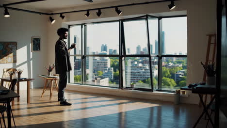 man drinking coffee and looking out a window in a bright art studio with a city view