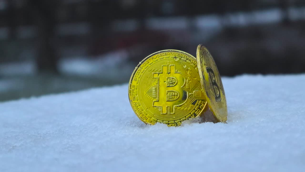 Camera moves around two golden yellow reflective Bitcoin coins while they stand up in white snow outdoors during winter time while it is snowing on and around the coins. Fallen snow is melting on coin