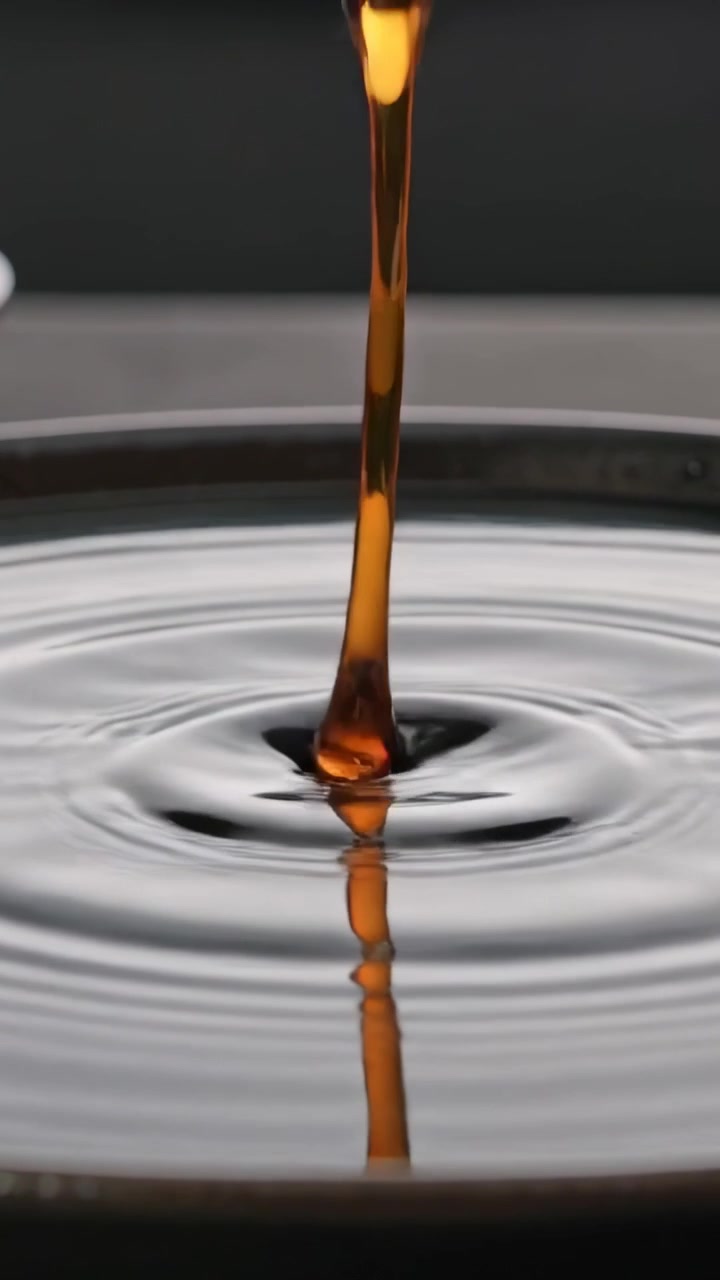 Vídeo vertical: Corrente âmbar sendo derramada, formando uma gota e atingindo uma tigela de estúdio, alimentando uma poça escura.
