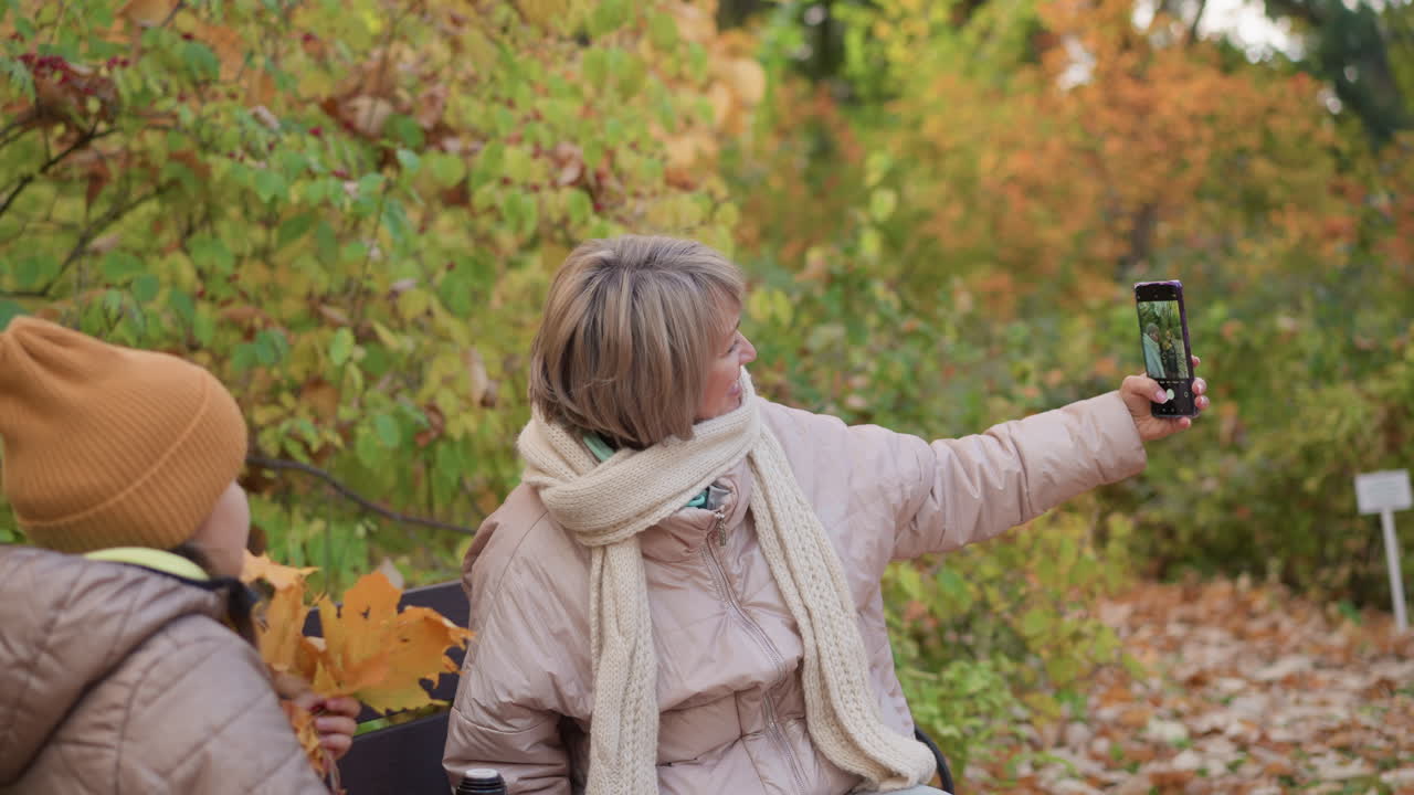 Mother wearing beige jacket and scarf stretches arm taking selfie with daughter holding autumn leaves while seated on bench surrounded by golden forest foliage on peaceful fall afternoon in nature park