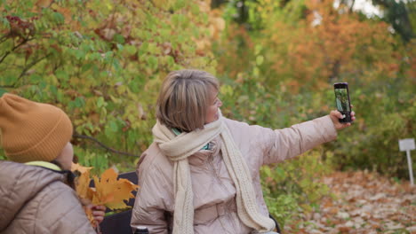 mother wearing beige jacket and scarf stretches arm taking selfie with daughter holding autumn leaves while seated on bench surrounded by golden forest foliage on peaceful fall afternoon in nature park