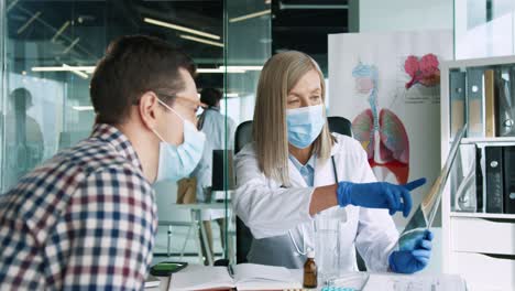 doctora caucásica con máscara médica sentada en el escritorio explicando la enfermedad pulmonar por coronavirus a un paciente masculino en consulta médica