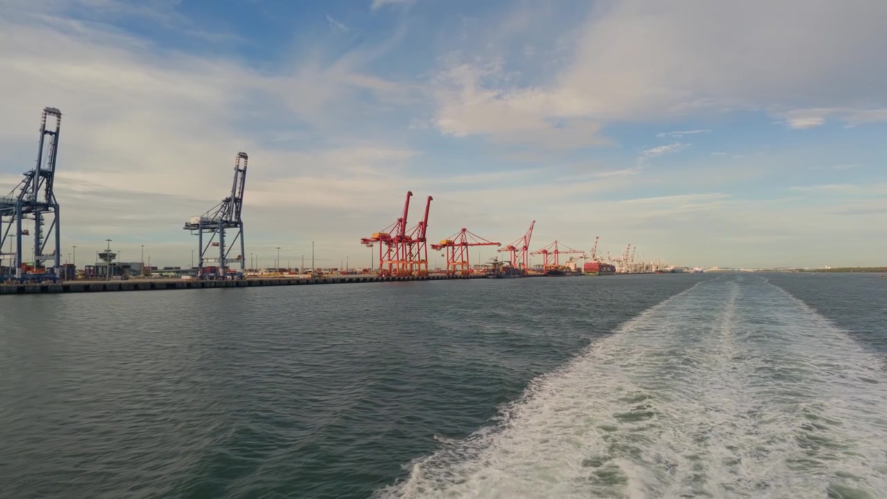 Mare calmo si estende davanti, scia che si trascina dietro. Grandi gru si ergono alte, porto industriale visibile. Cielo azzurro brillante, nuvole sparse delicatamente. La nave si muove costantemente attraverso le trafficate acque del porto.