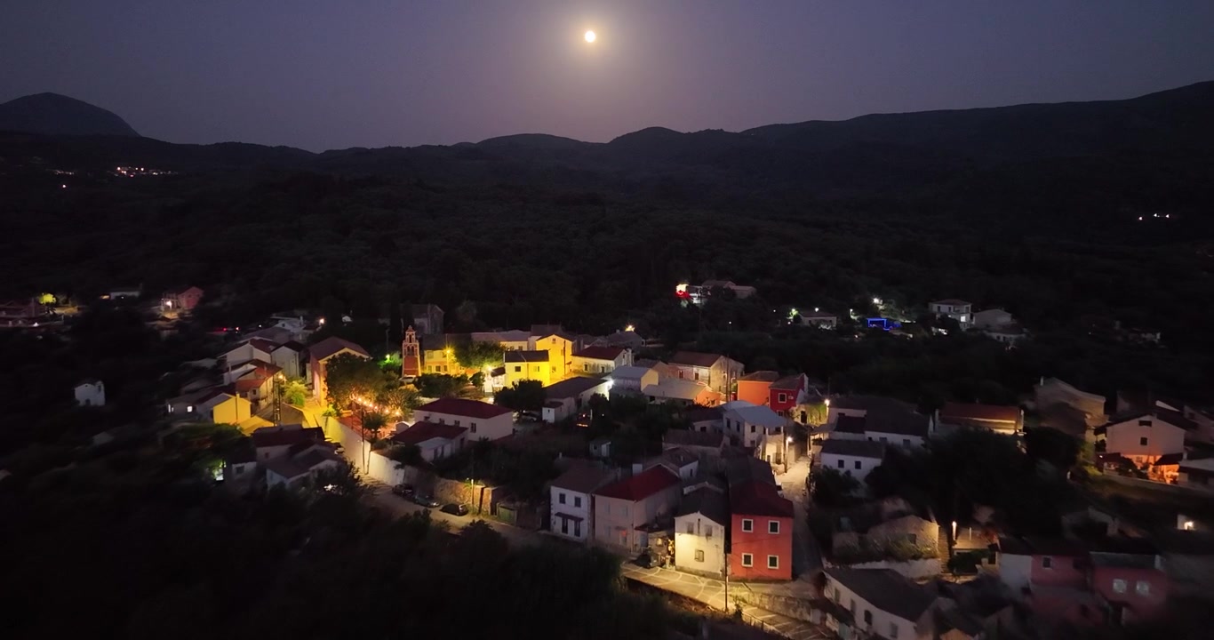 Luftaufnahme des Dorfzentrums Agioi Douloi auf der Insel Korfu, Griechenland, bei Nacht, mit leuchtenden Straßenlaternen, historischem Kirchturm unter hellem Vollmond