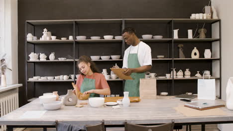 young woman wrapping ceramic bowl with paper while her male colleague writing on clipboard and then putting the bowl in a shopping bag at the pottery store