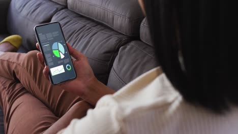 mixed race woman sitting on sofa using smartphone with statistics on screen