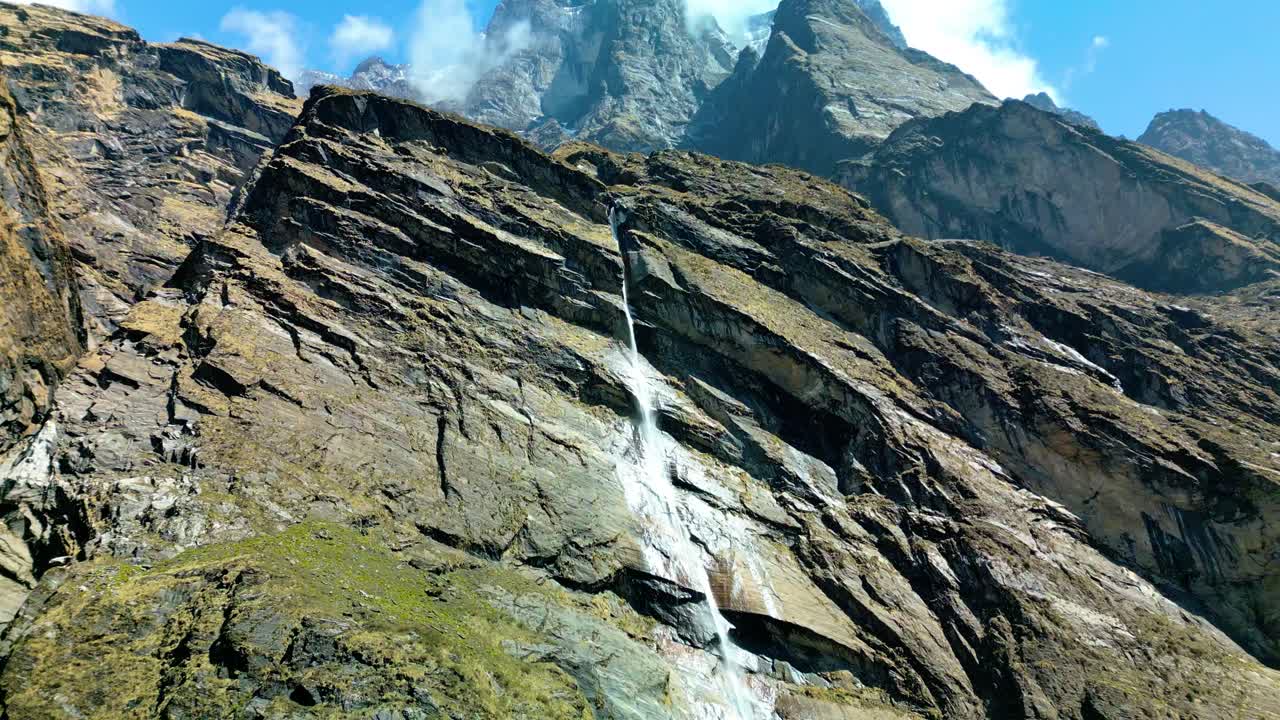 Beautiful waterfall cascading down a steep rocky mountain face in the Annapurna Conservation Area, surrounded by rugged cliffs, greenery, and clear blue Himalayan sky