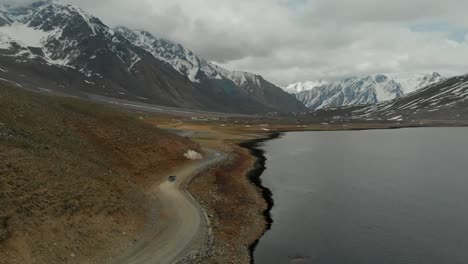 Vista-Aérea-De-La-Conducción-Suv-A-Lo-Largo-De-Un-Camino-Sinuoso-Al-Lado-Del-Lago-Shandur-En-Pakistán