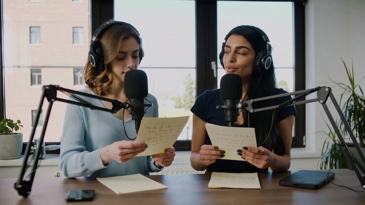 Recording starting hosts blue and dark tops reading scripts at desk window for podcast with mics