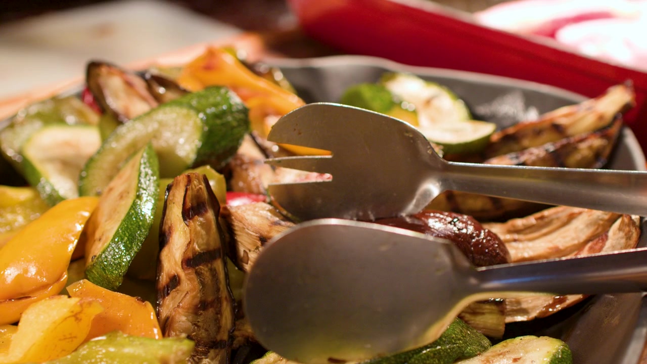 Pinze di metallo selezionano zucchine e peperoni grigliati da un vassoio di verdure colorate a un buffet.