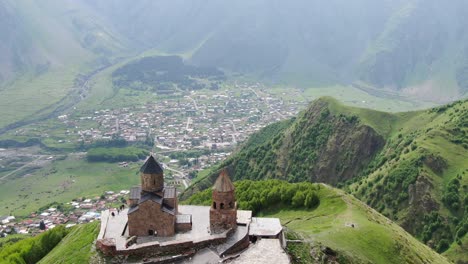 drone aerial view in georgia flying over gergeti trinity orthodox church and kazbegi town surrounded by green mountains valley with snowed peaks